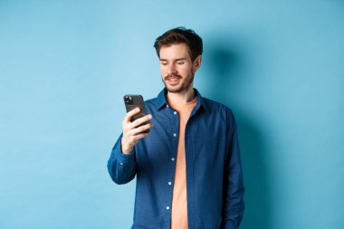 Cep telefonu kullanan yakışıklı, akıllı telefon ekranı okuyan ve gülümseyen, mavi arka planda iletişim kuran biri.