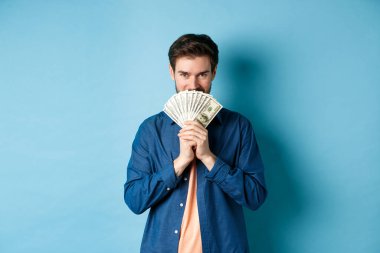 Genç adam, gülümseyen yüzünü paranın arkasına saklayıp kameraya bakarak mavi arka planda duruyor.