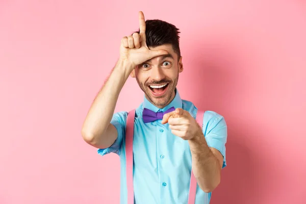 Imagen de tipo divertido apuntando a la cámara y mostrando signo perdedor, burlándose de la persona que pierde, de pie sobre el fondo rosa — Foto de Stock