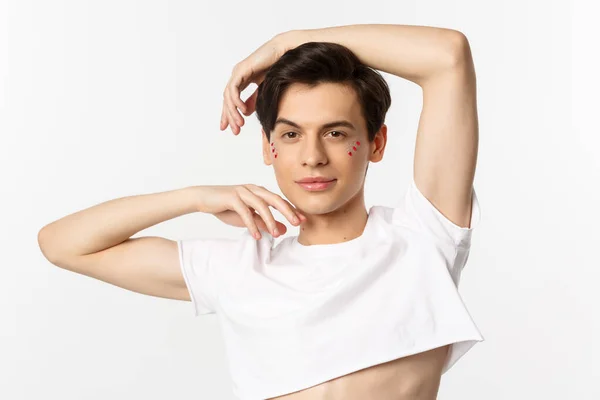 People, lgbtq community and lifestyle concept. Close-up of sassy gay man with lip gloss and glitter under eyes, posing for camera with pleased smile, wearing crop top, white background — Stock Photo, Image