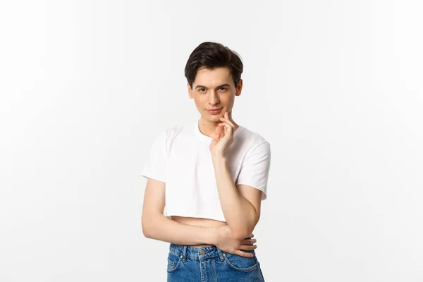 Thoughtful young queer man wearing crop top, smiling and looking at camera cunning, having an idea, standing over white background — Stock Photo, Image