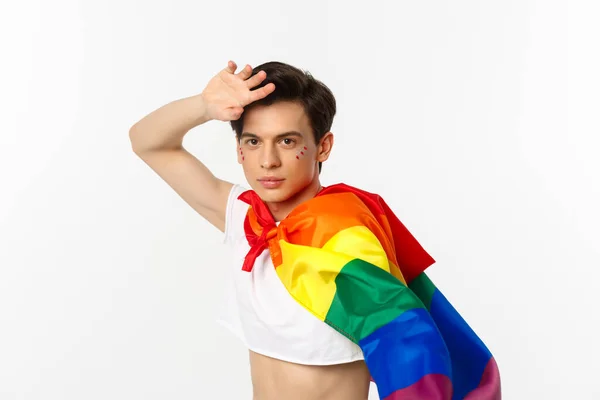 Hermoso hombre gay con brillo en la cara, vistiendo top de la cosecha y arco iris lgbt bandera, posando sobre fondo blanco —  Fotos de Stock
