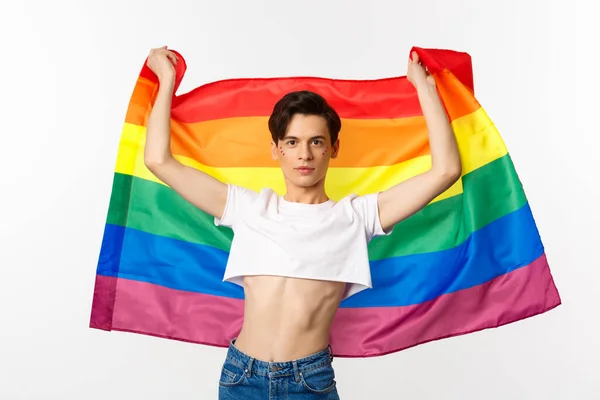 作物の上の若い男は、顔に輝き、自信を持って感情を誇りフラグを上げる。LGBTの旗で微笑むクィアの人 — ストック写真