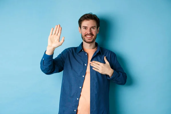 Joven feliz se presenta, diciendo la verdad con la mano en el corazón y la sonrisa amistosa, de pie sobre fondo azul — Foto de Stock