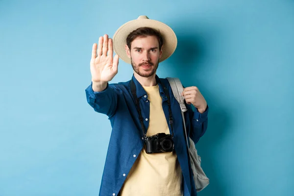 Serious male tourist warn you, stretch out hand to show stop gesture. Man on vacation forbid something, refusing or prohibiting action, standing with backpack on blue background