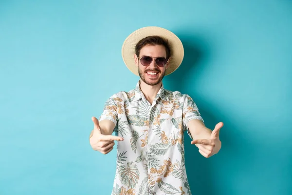 God turist i sommerhatt og hawaiisk skjorte, med logoen på midten, som viser noe, står på blå bakgrunn – stockfoto
