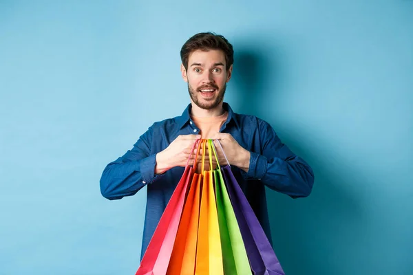 Feliz joven sosteniendo coloridas bolsas de compras y sonriendo emocionado, la compra de regalos, de pie sobre fondo azul —  Fotos de Stock