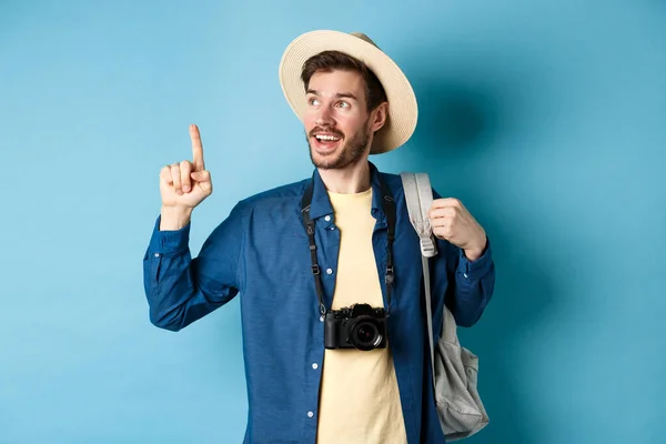 Bonito turista olhando e apontando para cima, viajando em férias de verão com mochila e câmera, de pé sobre fundo azul — Fotografia de Stock