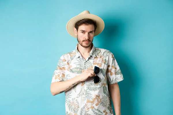 Kjekk turist i hawaiiskjorte og sommerhatt, legger solbriller i lomma, drar på ferie, står mot blå bakgrunn – stockfoto