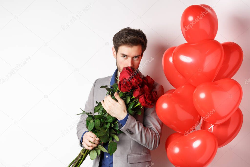 Romantic man smell bouquet of red roses and looking passionate at camera. Boyfriend in suit going on Valentines date with gifts and heart balloons, white background