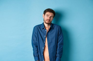 Gönülsüz ve üzgün bir adam üzgün hissediyor, boş bir yere bakıyor, güler yüzlü, mavi arka planda duruyor.