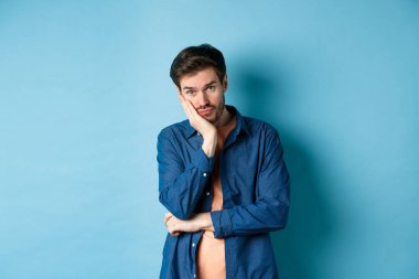 Üzgün ve sıkılmış bir adam elinde eğilmiş kameraya boş kafayla bakıyor, mavi arka planda sıkıntılı duruyor.