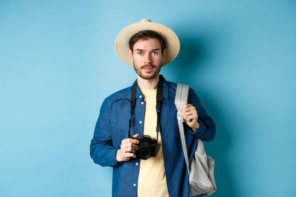 Kjekk ung turist i sommerhatt, ryggsekk og reise, med fotokamera stående på blå bakgrunn – stockfoto