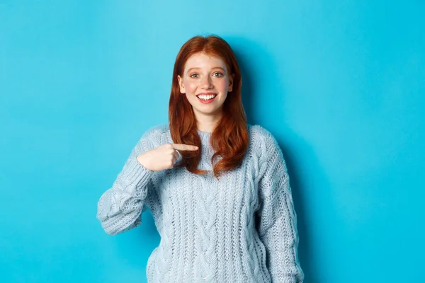 아름다운 빨간 머리 소녀가 자신을 가리키며 행복하게 웃고 있습니다. 파란 배경에 스웨터를 입고 — 스톡 사진