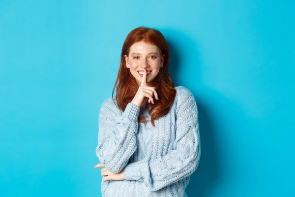 Adolescente ruiva bonita silenciando e sorrindo, contando um segredo, de pé em suéter contra fundo azul — Fotografia de Stock