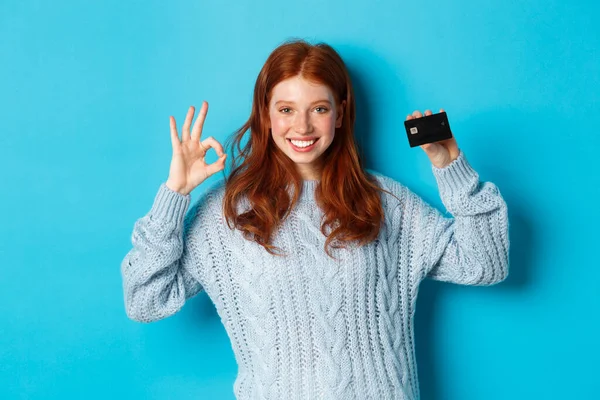 Happy redhead girl in sweater showing credit card and okay sign, recommending bank offer, standing over blue background