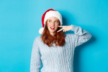 Kış tatili ve Noel arifesi konsepti. Kızıl saçlı mutlu genç kız, Noel Baba şapkası takıyor, yeni yılın tadını çıkarıyor, barış işareti gösteriyor ve tanıtıma soldan bakıyor, mavi arka planda duruyor.