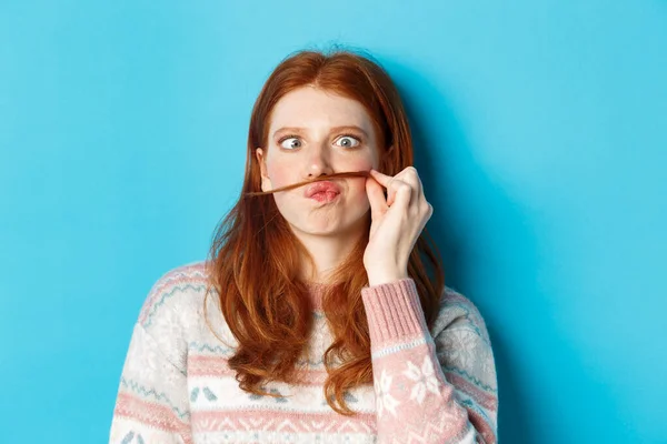 stock image Close-up of silly and funny redhead girl making moustache with hair strand and puckered lips, grimacing against blue background
