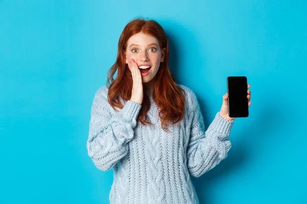 Удивительная рыжая девушка смотрит в камеру, показывает экран смартфона, демонстрирует мобильное приложение, стоя на синем фоне — стоковое фото