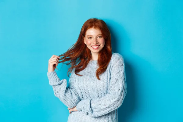 Flirtowa młoda kobieta z rudymi włosami, bawiąca się włosami i uśmiechnięta, stojąca w swetrze na niebieskim tle — Zdjęcie stockowe