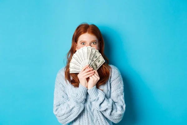 Mujer pelirroja emocionada cubriendo la cara con dinero, sosteniendo dólares y mirando a la cámara feliz, de pie sobre fondo azul — Foto de Stock