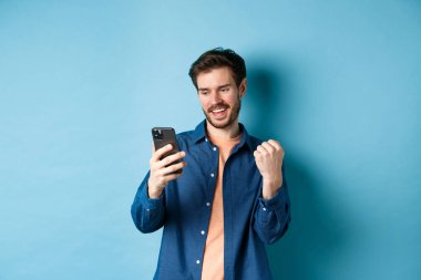 Şanslı adamın çevrimiçi kazandığı, cep telefonu ekranını okuduğu ve yumruk pompası yaptığı, zafer kazandığı, mavi arka planda durduğu görüntü