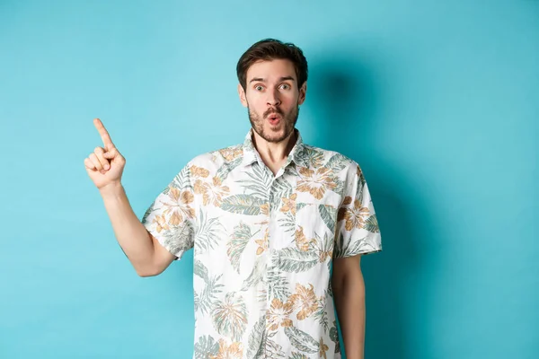 Sorprendido y sorprendido turista en camisa hawaiana apuntando con el dedo a la izquierda, mostrando promo y mirando fijamente a la cámara, de pie sobre fondo azul — Foto de Stock