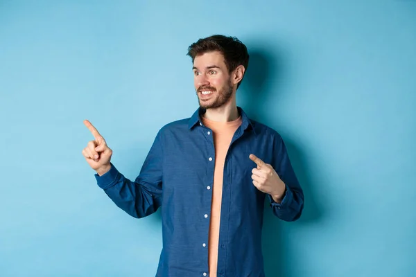 Sonriente chico guapo apuntando y mirando a la izquierda en el espacio vacío, mostrando el logotipo, de pie sobre fondo azul — Foto de Stock