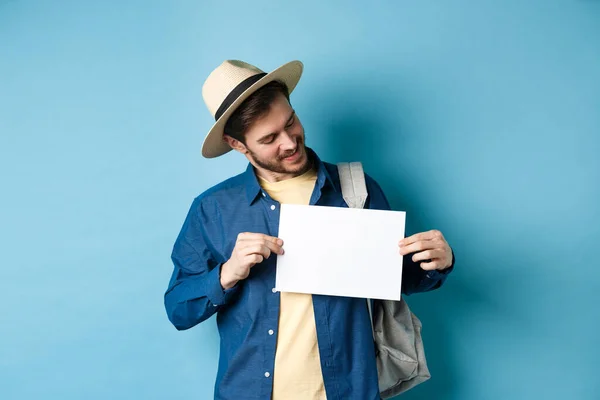 Turista feliz en sombrero de paja sonriendo y mirando un pedazo de papel para su logotipo, de pie sobre fondo azul — Foto de Stock