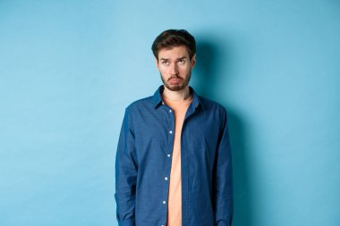 Üzgün adam mızmızlanıyor ve somurtuyor, üzgün bir suratla yukarı bakıyor, mavi arka planda duruyor.