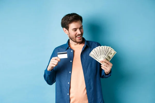 Sonriente chico moderno mirando en efectivo y mostrando tarjeta de crédito de plástico, de pie sobre fondo azul — Foto de Stock