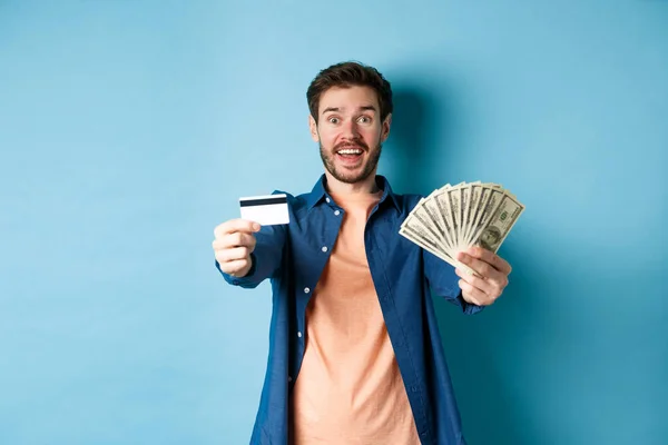 Chico alegre caucásico extendiendo las manos con dólares y tarjeta de crédito de plástico, sonriendo feliz en la cámara, de pie sobre fondo azul — Foto de Stock