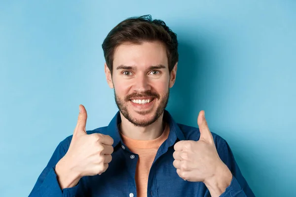 Close-up de sorrir homem bonito mostrando polegares para cima, elogiando bom trabalho, recomendando companhia, de pé sobre fundo azul — Fotografia de Stock