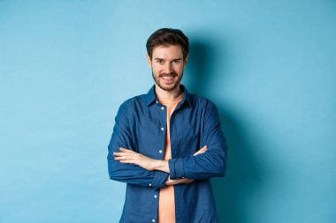 Kendine güveni tam genç bir adam kendini profesyonel hissediyor, kollarını göğsünde kavuşturuyor ve sırıtıyor, mavi arka planda duruyor.