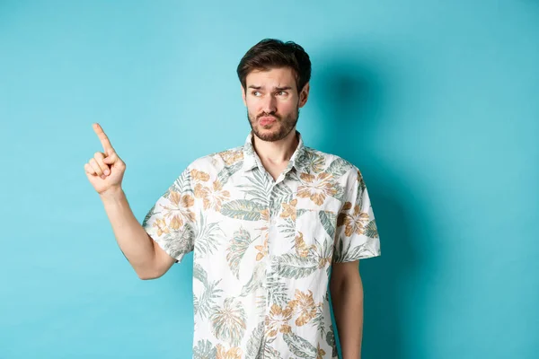 Δυσαρεστημένος τουρίστας με χαβανέζικο πουκάμισο, κοιτάζοντας και δείχνοντας αριστερά απογοητευμένος, παραπονιέται για κάτι κακό, στέκεται σε μπλε φόντο — Φωτογραφία Αρχείου