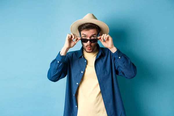 Retrato de turista decolagem óculos de sol e dizendo uau, olhando impressionado para a câmera, verificando coisa legal, de pé em chapéu de verão no fundo azul — Fotografia de Stock