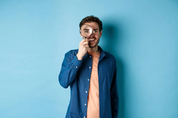 Cheerful young man found something good, smiling and looking through magnifying glass, standing on blue background — Stock Photo, Image