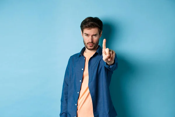 Serious young man extend one finger to stop or warn you, prohibit something or disapprove, standing on blue background — Stock Photo, Image