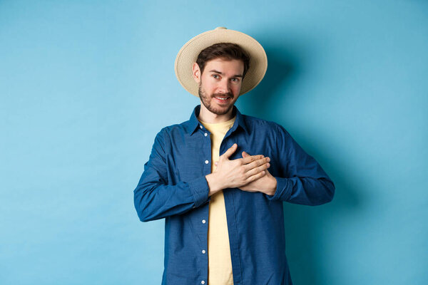 Handsome traveler in straw hat, holding hands on heart and smiling, showing thank you gesture, keep memories about vacation in soul, standing on blue background