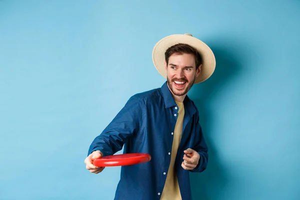 Hombre feliz riendo y jugando frisbee, lanzando a amigo y sonriendo, de pie en sombrero de verano sobre fondo azul — Foto de Stock