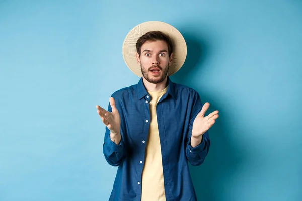 Geschokte mannelijke toerist op vakantie handen omhoog en kijken naar camera geschrokken, reageren op groot nieuws met ongeloof, staan op blauwe achtergrond in stro zomer hoed en shirt — Stockfoto