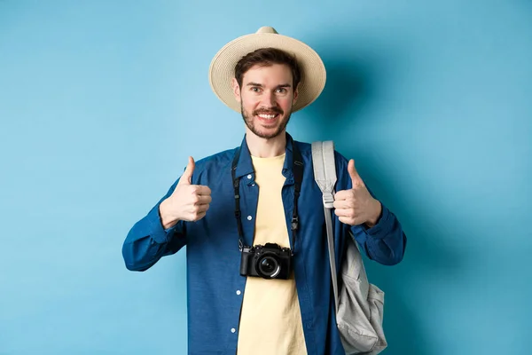 Homem bonito alegre recomendando lugar de férias de verão, mostrando polegares para cima e sorrindo na aprovação. Turista deixar feedback positivo, de pé com câmera e mochila no fundo azul — Fotografia de Stock