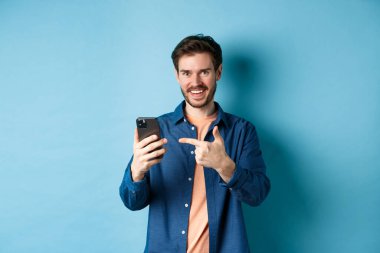 Yakışıklı gülümseyen adam akıllı telefonu işaret ediyor ve kameraya memnun bakıyor, uygulamayı tavsiye ediyor, mavi arka planda duruyor.