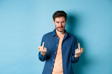 Ignorant and unbothered man showing middle-fingers and looking reluctant at camera, give zero fucks, standing on blue background
