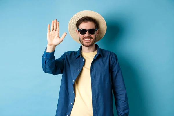 Kjekk hipster på sommerferie, gir avkall på hånden og smiler, sier hallo, bruker solbriller og stråhatt, blå bakgrunn – stockfoto