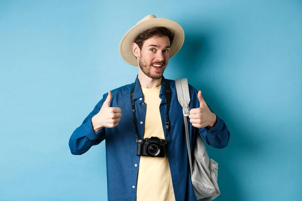 Feliz turista sorridente dizer sim e mostrar polegares para cima, indo de férias de verão, segurando mochila e câmera de foto, fundo azul — Fotografia de Stock