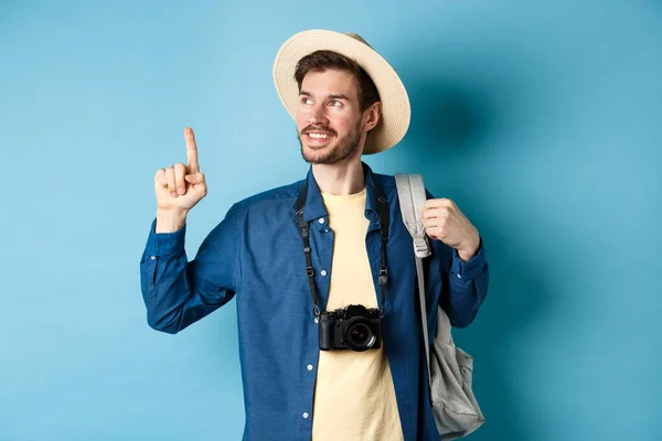 Bonito cara positivo em férias, usando chapéu de verão, olhando e apontando para o logotipo, sorrindo satisfeito, de pé com câmera e mochila no fundo azul — Fotografia de Stock