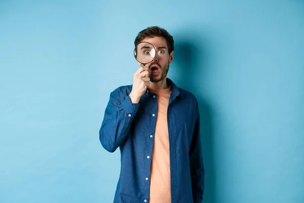Indrukwekkende man kijken in ontzag door vergrootglas, vallen kaak en staren naar de camera, staan op een blauwe achtergrond — Stockfoto