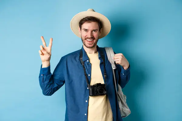 Lykkelig, kjekk mann som tar bilde på sommerferie, viser fredskilt og smiler, går med stråhatt og holder turistryggsekk, blå bakgrunn – stockfoto