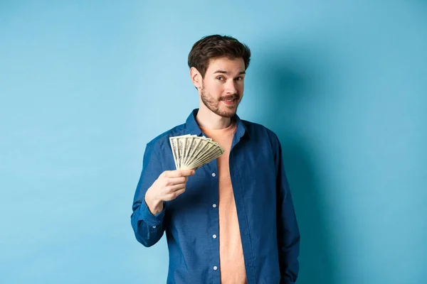 Tipo rico sin inconvenientes mostrando dinero en efectivo y sonriendo, sosteniendo dólares en la mano, de pie sobre fondo azul — Foto de Stock